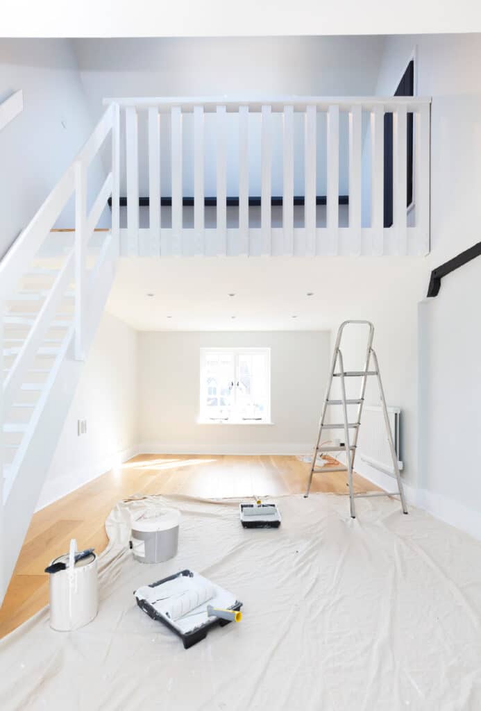 Intérieur blanc en rénovation: escalier, mezzanine, sol bois. Peinture, pinceaux, rouleau et échelle sur une bâche.