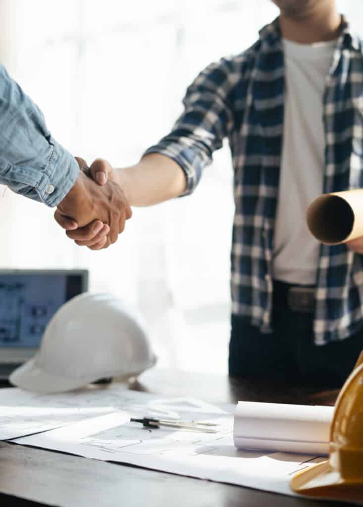 Deux hommes, probablement architectes ou ingénieurs, se serrent la main sur un bureau avec des plans, un ordinateur et un casque. Symbole de collaboration.